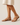 "Woman wearing beige flat leather sandals with ankle strap and gold buckle, styled with a cream midi dress, name-brand summer footwear modeled against a neutral background."