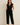 "Woman wearing a black sleeveless belted jumpsuit with tan sandals, name-brand women’s jumpsuit modeled for casual and elegant fashion against neutral background."