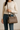 Woman wearing a cream ribbed sweater and blue jeans holding a taupe leather crossbody handbag, name-brand women's purse modeled for everyday fashion and functional style against neutral background.